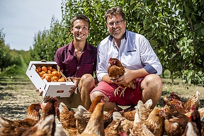 Zwei Personen sitzen von Hühnern umgeben in einem Garten. Eine Person hält eine Kiste mit Zitrusfrüchten. © Netzwerk Kulinarik / pov.at
